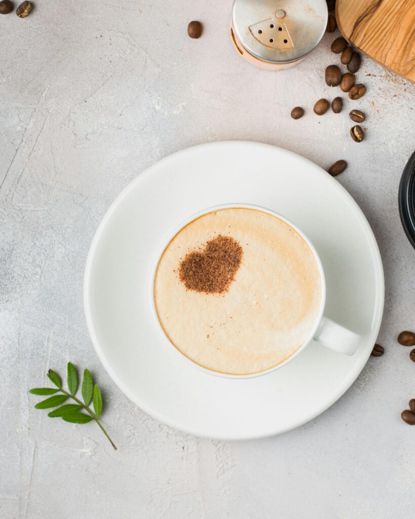 A cappuccino with a heart-shaped cocoa dusting on a saucer, surrounded by coffee beans.