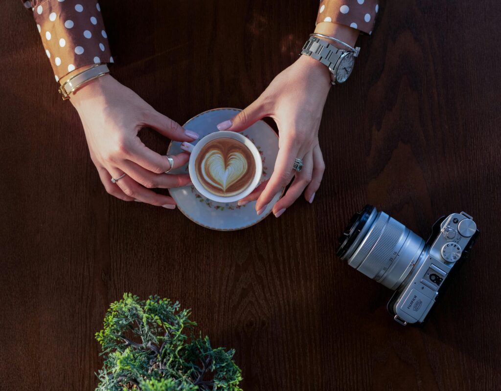 Hands holding a coffee cup with latte art next to a retro camera on a wooden table.